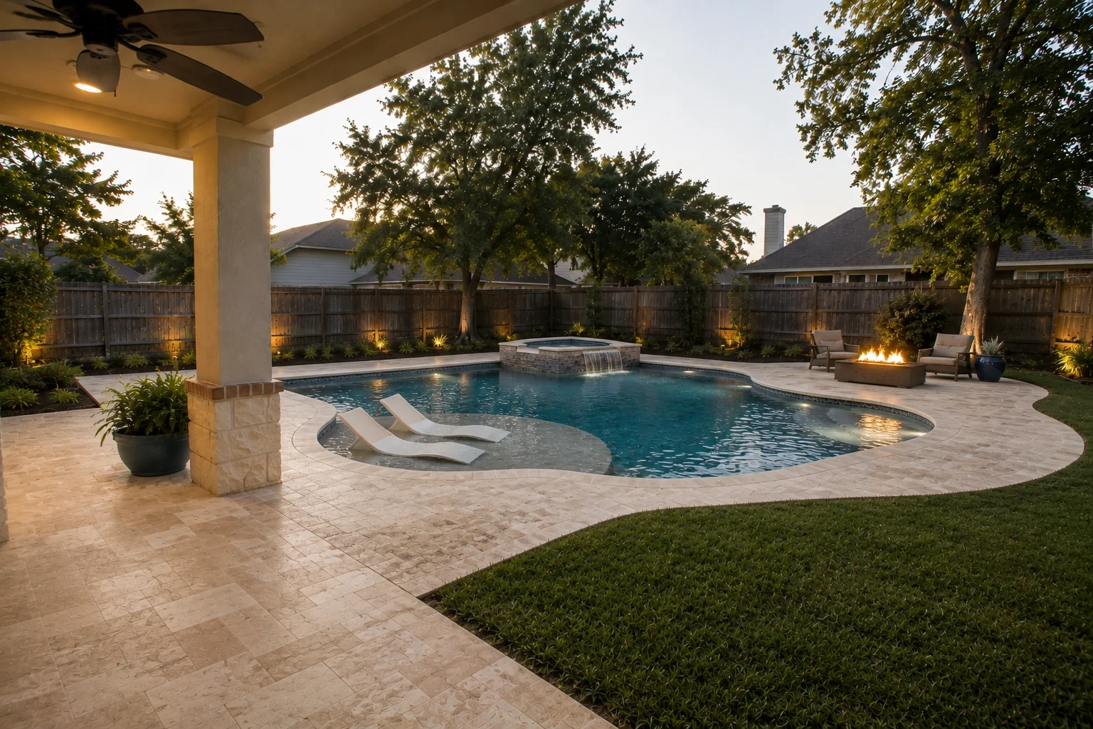 The same yard transformed at dusk: flowing soft-geometric pool with wide tanning ledge and in-water loungers, raised stone spa with spillway, travertine deck curving with the shell, fire-pit seating, perimeter beds uplit, generous lawn retained