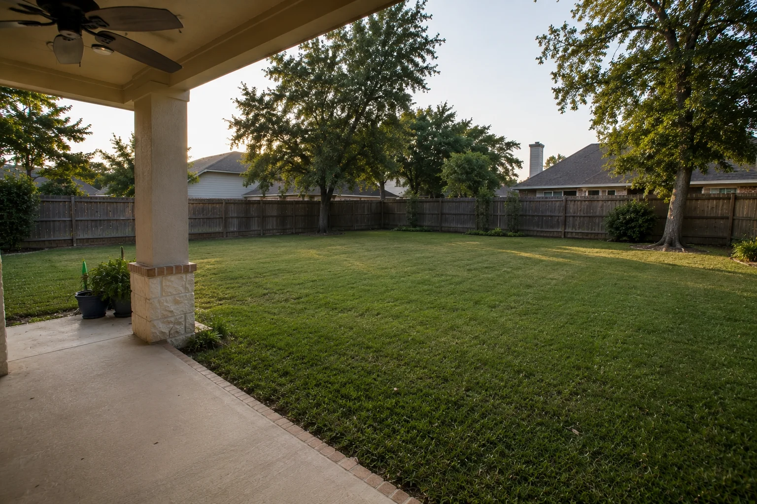 Plain fenced Houston family backyard from a covered patio with stone-based column: open lawn, mature trees, neighboring rooflines and chimney