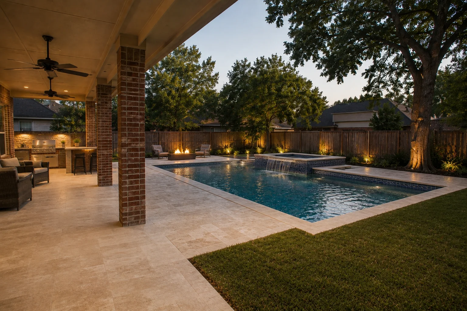 The same yard transformed at dusk: geometric pool with raised spillover spa, expanded travertine deck, outdoor kitchen with bar seating, fire-pit lounge by the fence, the same oak uplit in a planted bed, lawn retained