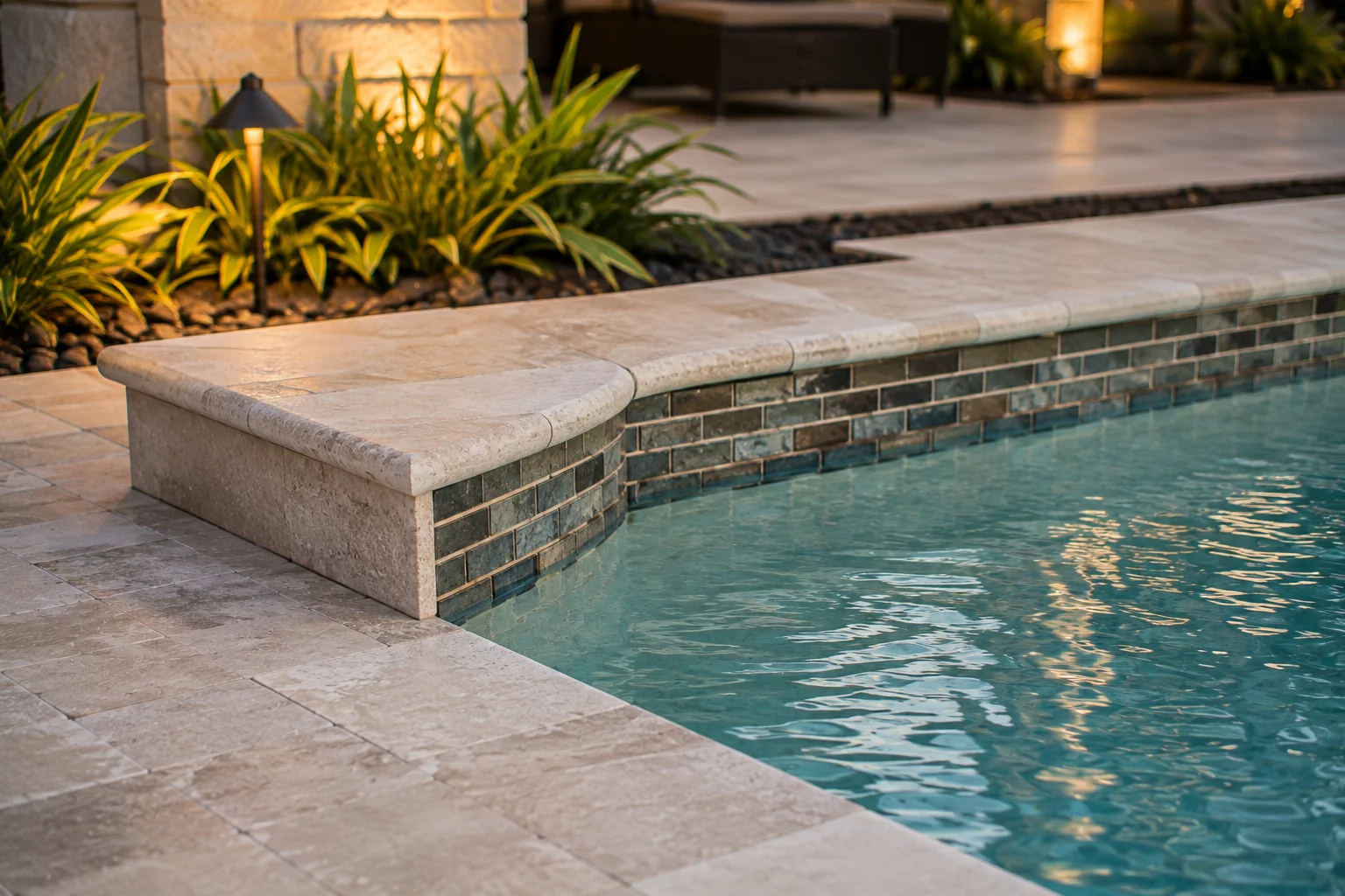 Close detail of rounded travertine coping over glass-and-slate waterline tile, with river-stone border and lit planting beyond
