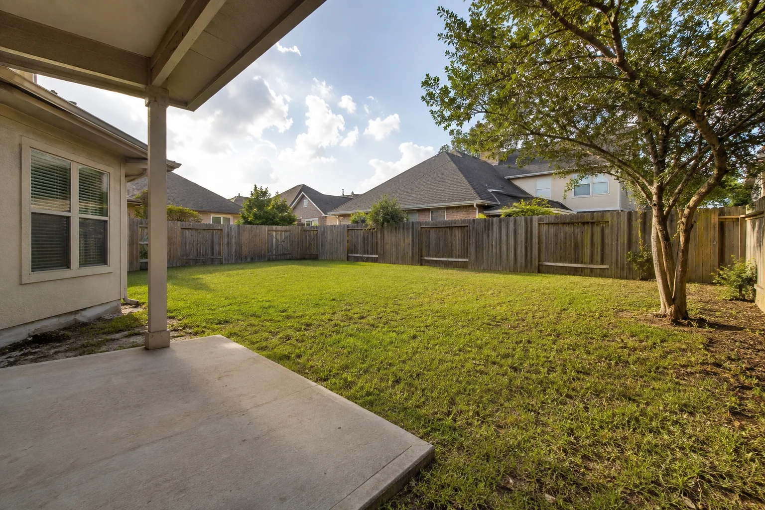 Plain Houston backyard from a small covered porch: bare concrete slab, patchy lawn, weathered privacy fence with gates, multi-trunk crepe myrtle, neighboring rooflines