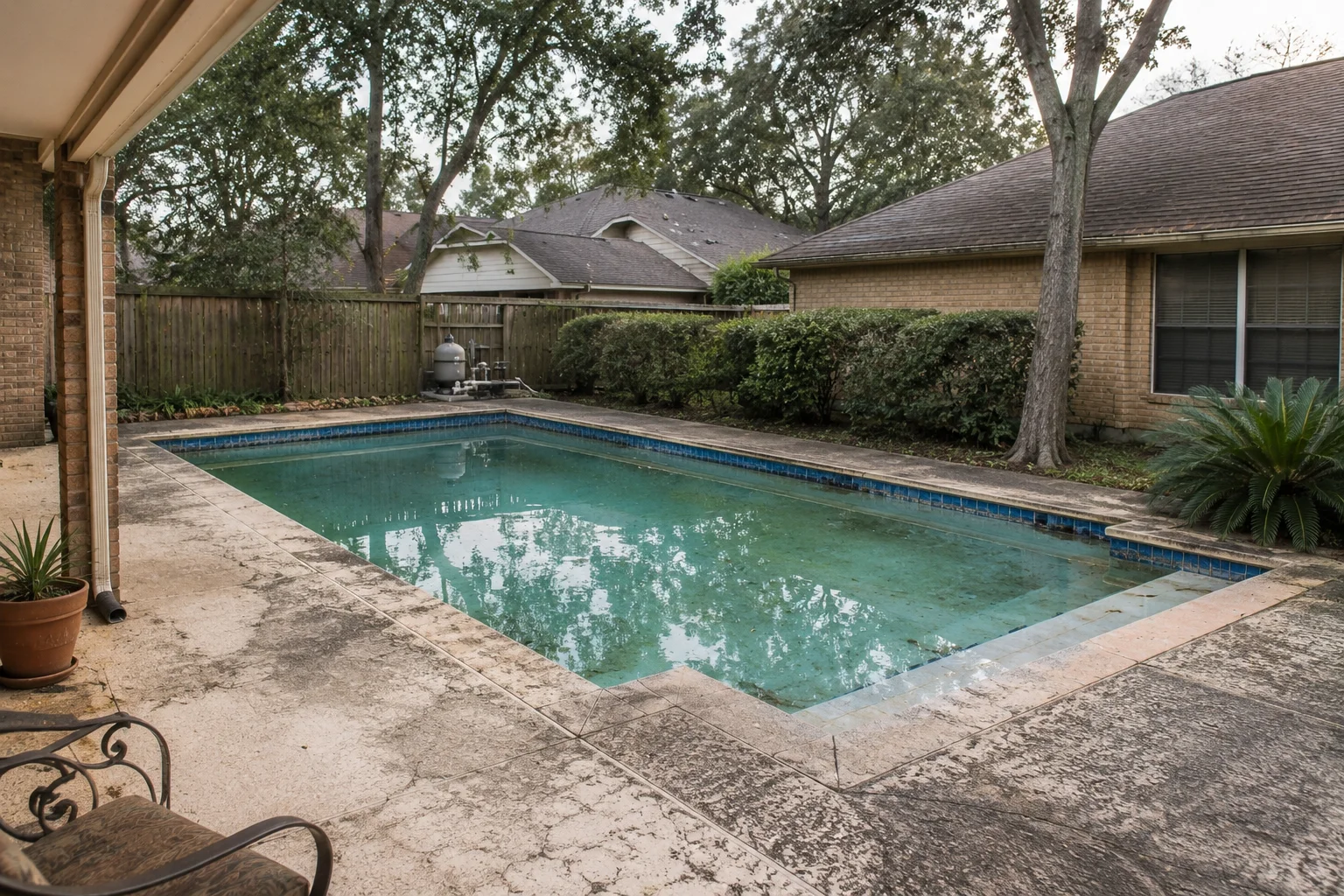 Aging backyard pool with faded mottled water, dated blue waterline tile, stained weathered concrete deck, and tired plantings beside a brick ranch home